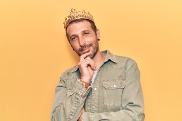 Young handsome man wearing king crown smiling looking confident at the camera with crossed arms and hand on chin. thinking positive.