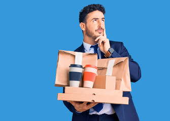 Young hispanic man wearing suit holding takeaway coffee and food serious face thinking about...
