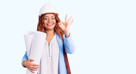 Young latin woman wearing architect hardhat and leather bag holding blueprints doing ok sign with fingers, smiling friendly gesturing excellent symbol
