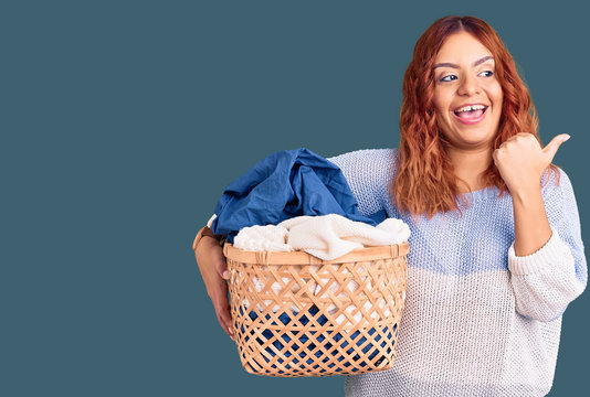Young latin woman holding laundry basket pointing thumb up to the side smiling happy with open mouth