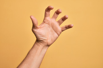 Hand of caucasian young man showing fingers over isolated yellow background grasping aggressive and scary with fingers, violence and frustration