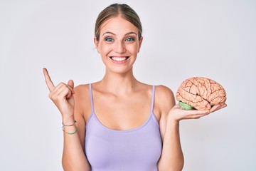 Young blonde girl holding brain smiling happy pointing with hand and finger to the side