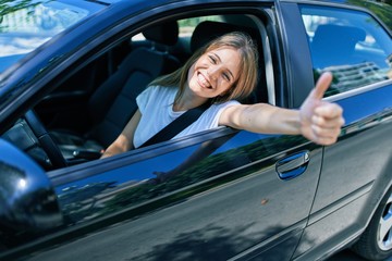 Young beautiful blonde woman smiling happy with thumb up doing ok sign sitting at the car