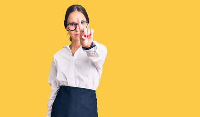 Beautiful brunette young woman wearing professional waitress apron pointing with finger up and angry expression, showing no gesture