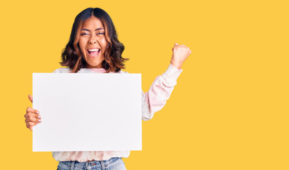 Fototapeta premium Young beautiful mixed race woman holding cardboard banner with blank space screaming proud, celebrating victory and success very excited with raised arms