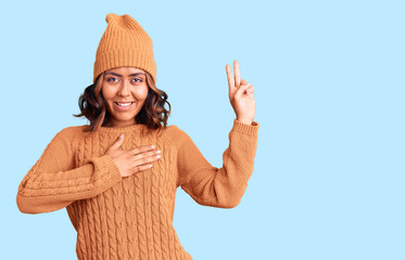 Young beautiful mixed race woman wearing wool sweater and winter hat smiling swearing with hand on...