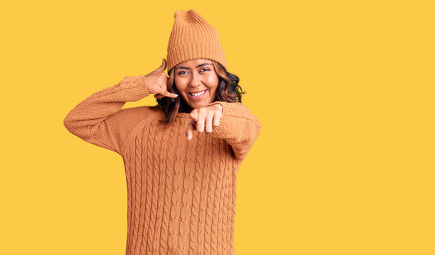 Young beautiful mixed race woman wearing wool sweater and winter hat smiling doing talking on the telephone gesture and pointing to you. call me.