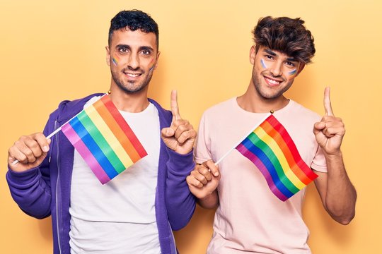 Young gay couple holding rainbow lgbtq flags smiling with an idea or question pointing finger with happy face, number one