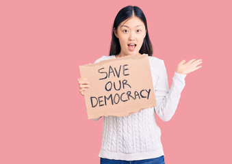 Young beautiful chinese woman holding save our democracy cardboard banner celebrating victory with happy smile and winner expression with raised hands