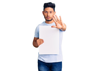 Handsome latin american young man holding cardboard banner with blank space with open hand doing stop sign with serious and confident expression, defense gesture