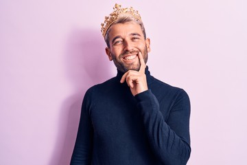 Young handsome blond man with piercing wearing golden king crown over pink background smiling looking confident at the camera with crossed arms and hand on chin. Thinking positive.