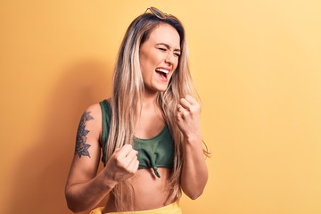 Young beautiful blonde woman wearing bikini and sunglasses standing over yellow background celebrating surprised and amazed for success with arms raised and eyes closed