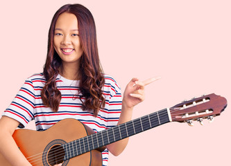 Fototapeta premium Young beautiful chinese girl playing classical guitar smiling happy pointing with hand and finger to the side