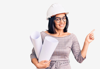 Young beautiful latin woman wearing architect hardhat and glasses holding blueprints smiling happy pointing with hand and finger to the side