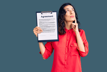 Young beautiful hispanic woman holding clipboard with agreement document serious face thinking about question with hand on chin, thoughtful about confusing idea