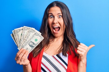 Beautiful brunette businesswoman holding bunch of dollars banknotes over blue background pointing thumb up to the side smiling happy with open mouth