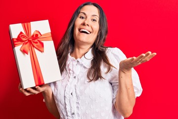 Young beautiful brunette woman holding birthday gift standing over isolated red background celebrating achievement with happy smile and winner expression with raised hand