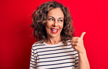 Middle age beautiful brunette woman wearing striped t-shirt standing over red background pointing thumb up to the side smiling happy with open mouth