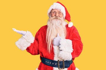 Old senior man with grey hair and long beard wearing traditional santa claus costume showing palm hand and doing ok gesture with thumbs up, smiling happy and cheerful
