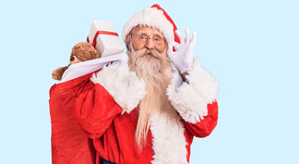 Old senior man with grey hair and long beard wearing santa claus costume holding bag with presents doing ok sign with fingers, smiling friendly gesturing excellent symbol