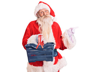 Old senior man with grey hair and long beard wearing santa claus costume holding shopping basket smiling happy pointing with hand and finger to the side