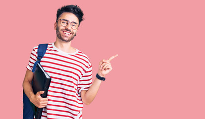 Young hispanic man wearing student backpack and glasses holding binder smiling happy pointing with hand and finger to the side