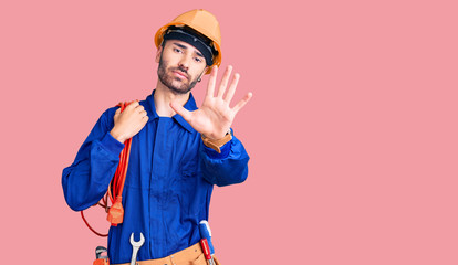 Young hispanic man wearing elecrician uniform holding cable with open hand doing stop sign with serious and confident expression, defense gesture