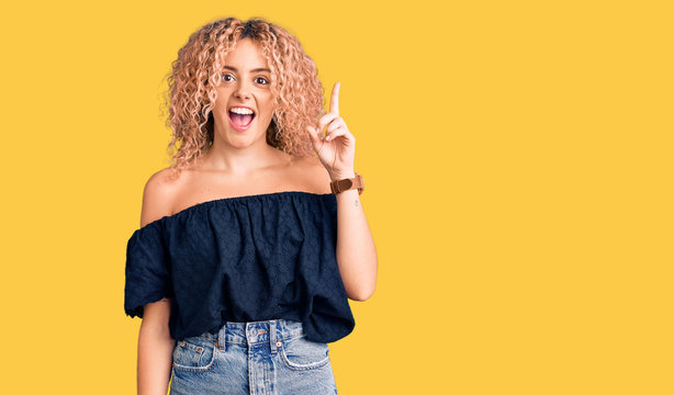 Young blonde woman with curly hair wearing casual summer clothes pointing finger up with successful idea. exited and happy. number one.