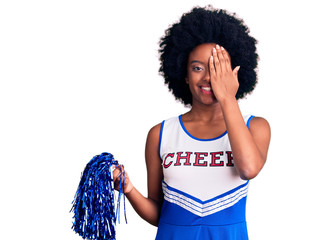 Young african american woman wearing cheerleader uniform holding pompom covering one eye with hand,...