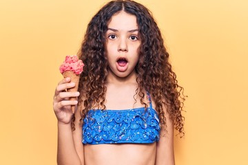 Beautiful kid girl with curly hair wearing bikini holding ice cream scared and amazed with open...