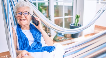 Elder senior woman with grey hair smiling happy relaxing on a hammock at home speaking on the phone