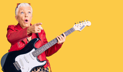 Fototapeta premium Senior beautiful woman with blue eyes and grey hair wearing a modern look playing electric guitar pointing with finger surprised ahead, open mouth amazed expression, something on the front