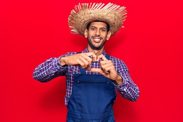 Young latin man wearing farmer hat and apron smiling in love doing heart symbol shape with hands. romantic concept.
