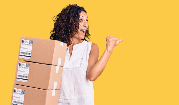 Middle age beautiful woman holding delivery package pointing thumb up to the side smiling happy with open mouth