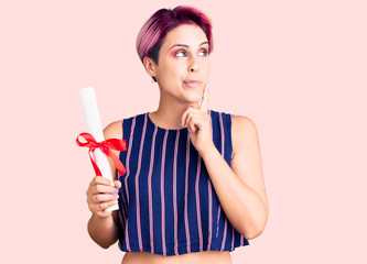 Young beautiful woman with pink hair holding graduate degree diploma serious face thinking about question with hand on chin, thoughtful about confusing idea