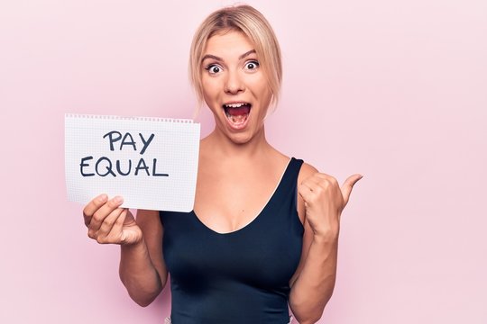 Beautiful blonde woman asking for equality economy holding paper with pay equal message pointing thumb up to the side smiling happy with open mouth