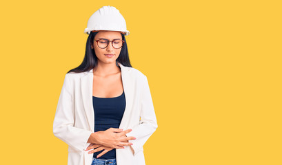 Young beautiful latin girl wearing architect hardhat and glasses with hand on stomach because indigestion, painful illness feeling unwell. ache concept.