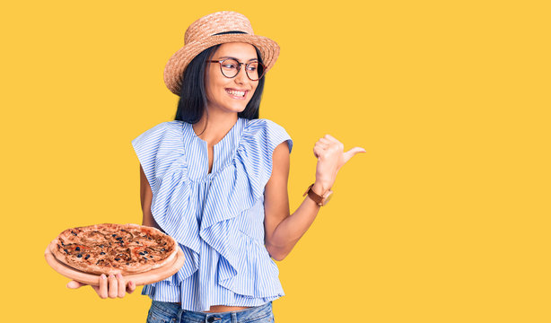Young beautiful latin girl wearing summer hat and glasses holding pizza pointing thumb up to the side smiling happy with open mouth