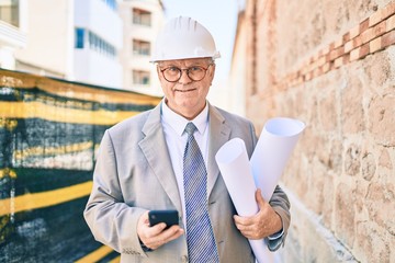 Senior grey-haired architect man holding blueprints using smartphone at street of city