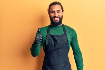 Fototapeta premium Young arab man wearing barber apron smiling happy and positive, thumb up doing excellent and approval sign
