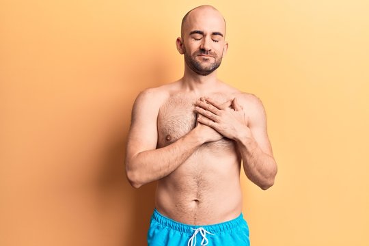 Young handsome bald man wearing swimwear shirtless smiling with hands on chest, eyes closed with grateful gesture on face. health concept.