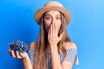 Young beautiful girl wearing summer hat holding bowl with blueberries covering mouth with hand, shocked and afraid for mistake. surprised expression