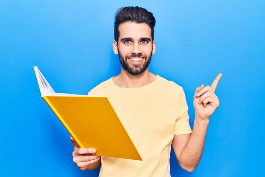 Young handsome man with beard reading book smiling happy pointing with hand and finger to the side