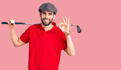 Young handsome man with beard playing golf holding club and ball doing ok sign with fingers, smiling friendly gesturing excellent symbol