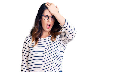 Beautiful young brunette woman wearing casual clothes and glasses surprised with hand on head for...