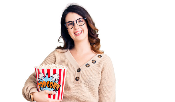 Beautiful young brunette woman eating popcorn looking positive and happy standing and smiling with a confident smile showing teeth