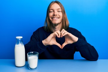 Handsome caucasian man with long hair holding glass of milk smiling in love doing heart symbol shape with hands. romantic concept.