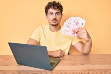 Young caucasian man with curly hair sitting on the table working with laptop and holding israel shekels banknotes thinking attitude and sober expression looking self confident