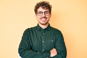 Young caucasian man with curly hair wearing casual clothes and glasses happy face smiling with crossed arms looking at the camera. positive person.