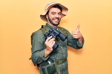 Young hispanic man wearing explorer clothes holding binoculars smiling happy and positive, thumb up doing excellent and approval sign